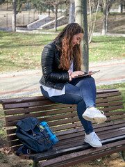 Young caucasian female using her tablet while sitting on a wooden bench in the park