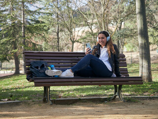 Young female with headphones using her smartphone while sitting on a wooden bench in the park
