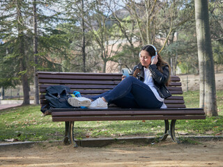 Young female with headphones using her smartphone while sitting on a wooden bench in the park
