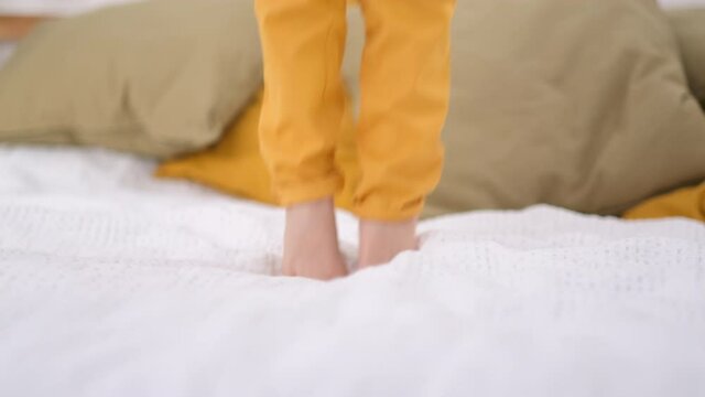 Close up legs jumping on white bed. Excited caucasian boy in yellow pants having fun at home enjoy laughing playing funny active game in bedroom.