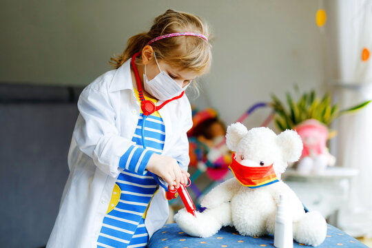 Little Girl Makes Injection To Teddy Bear. Cute Child With Medical Mask Playing Doctor, Holding Syringe With Vaccine. Coronavirus Covid Vaccination Concept. Kid Play Role Game At Home.