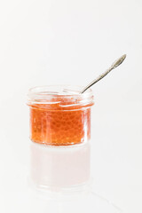 Glass jar with red salmon caviar fish on a white background with reflection . The jar is open, there is a spoon inside.