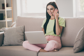 Photo of charming girl sit couch hold phone conversation laptop look screen wear glasses green t-shirt pink pants indoors