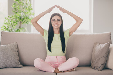 Fototapeta na wymiar Photo of positive young lady sit couch crossed legs hands above head roof shape wear glasses green t-shirt pink pants indoors