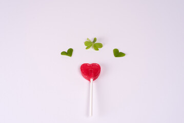 Red heart shaped lollipop with clover leaves flying above, on a light pink background. Concept of love, Valentine