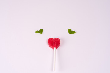 Red heart shaped lollipop with clover leaves flying above, on a light pink background. Concept of love, Valentine 
