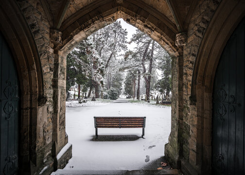 Winter churchyard grave yard stones solitary single bench in falling snow winter scene church archway beautiful pathway peaceful blanket of snowfall Nottinghamshire cemetery local peaceful thought