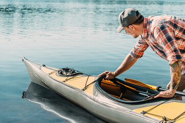 Young man in cap and shirt pushes a kayak in the water of lake on a summer sunny day.