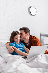 happy father kissing cheerful daughter using laptop in hospital bed