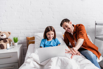 cheerful father pointing with fingers at laptop near happy daughter in hospital bed