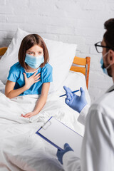 child in medical mask touching chest while talking to doctor with clipboard on blurred foreground