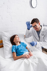 Obraz premium cheerful pediatrician gesturing and sticking out tongue while holding thermometer near happy girl lying in bed