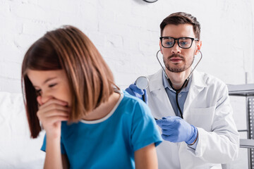 doctor in eyeglasses examining coughing girl with stethoscope in clinic, blurred foreground