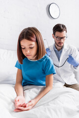 diseased girl sitting on bed in clinic near pediatrician with stethoscope on blurred background