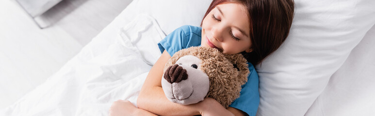 top view of smiling girl embracing teddy bear while sleeping in clinic, banner