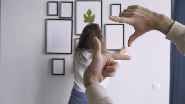 Person holds fingers in photo frame form against blurred lady with long loose curly hair decorating wall with pictures of different size in new flat