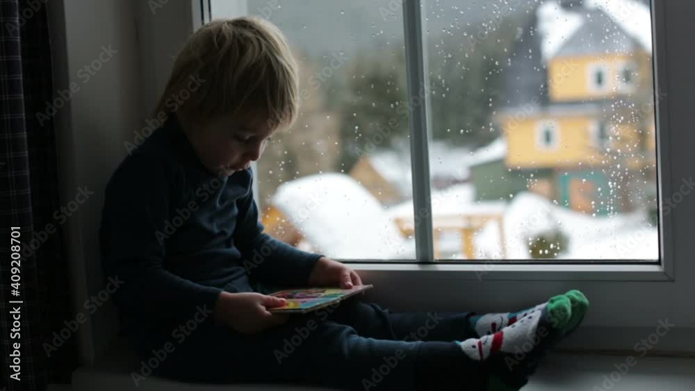 Canvas Prints Toddler child, sitting on the window, watching the snow falling, reading little book