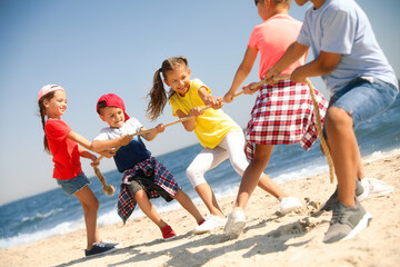 Cute children pulling rope during tug of war game on beach. Summer camp - obrazy, fototapety, plakaty
