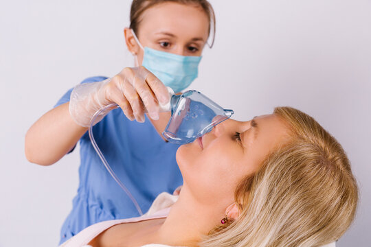 Girl Doctor In A Medical Uniform And In A Mask Holds A Nebulizer To An Older Woman On A White Background. Treatment Of Colds. Enlarged Photo.