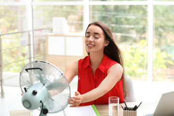 Young woman enjoying air flow from fan at workplace. Summer heat