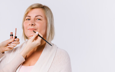 Lip contour and lipstick. Hands of the master do gentle makeup for an older woman on a white background with empty side space.