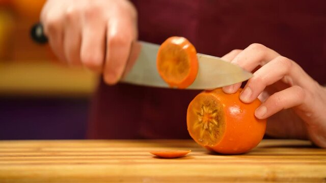 Man's hand cutting Persimmon kinglet orange.  Male's hand cutting fruits with sharp knife in kitchen. Man is slicing orange ripe persimmon  fruit, close view.  