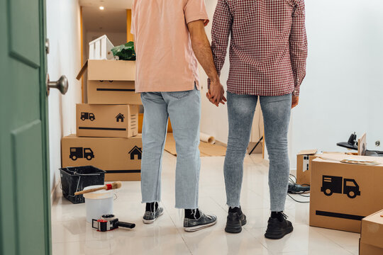 Unrecognizable Gay couple, from the back, holding hands standing at their new home together. Cropped image of a couple in love moving at new apartment, with pile of cardboard boxes on the floor.