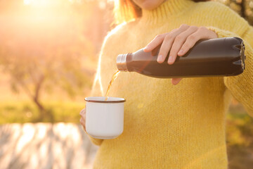 Woman pouring hot drink from thermos bottle into cup outdoors, closeup - obrazy, fototapety, plakaty
