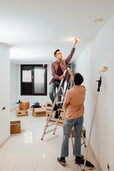 Young man on top of a stairway fixing a light bulb while his friend helps to hold the ladder. Gay couple doing renovations at a new home, apartment, with cardboard boxes on floor.