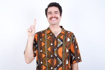Young caucasian man wearing generic pattern printed shirt against yellow wall showing and pointing up with finger number one while smiling confident and happy.