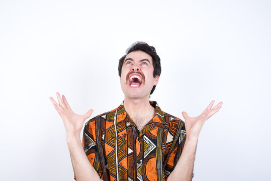 Young caucasian man wearing generic pattern printed shirt against yellow wall crazy and mad shouting and yelling with aggressive expression and arms raised. Frustration concept.