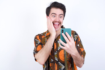 Young caucasian man wearing generic pattern printed shirt against white wall holding in hands cell reading sms using new app 5g