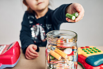 Little girl putting used batteries into jar for recycling. Child separating waste. Batteries only container - obrazy, fototapety, plakaty