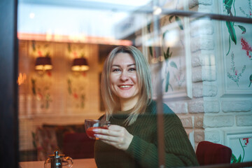 Woman in sweater sitting in restaurant with cup of fruit tea and looking out the window, view through glass