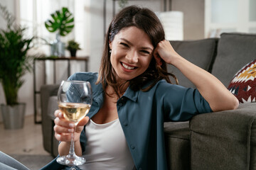 Happy woman sitting on the floor, drinking wine. Smiling woman celebrate with wine at home..
