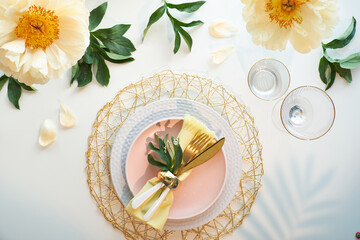 Fototapeta na wymiar Effortless golden birthday dinner table decor. Pale yellow peony flowers and Late Spring, Summer flat lay. White dinner table, white and gold utensils, decorated with peony flowers.