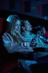 Beautiful young woman smiling, using her phone while missing boring movie at the cinema