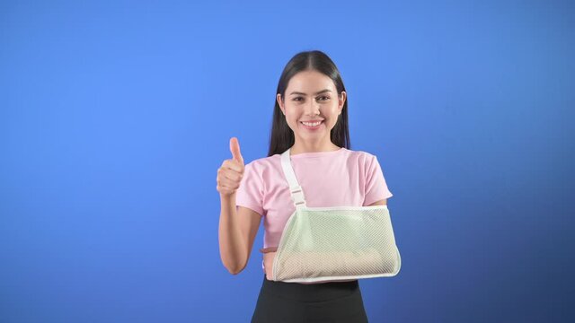 Portrait of young woman with an injured arm in a sling over blue background in studio, insurance and healthcare concept