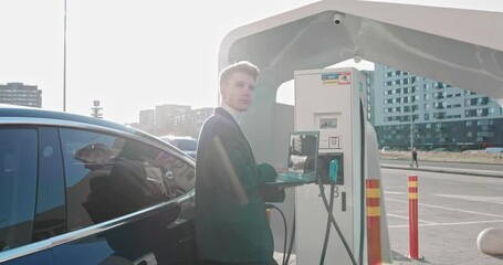 Businessman holds a laptop in his hands and leans on a blue luxury electric car, which is charging. Work While Charging The Car. A luxury electric car is charged at an electric charging station