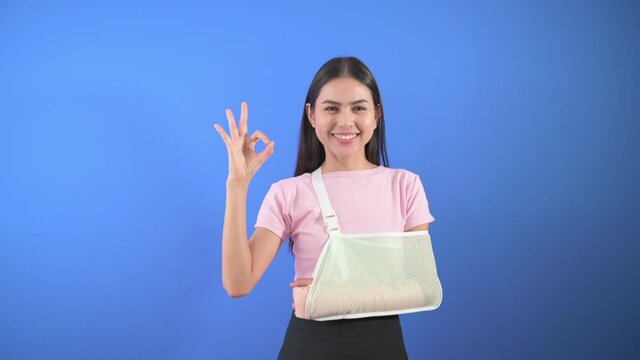 Portrait of young woman with an injured arm in a sling over blue background in studio, insurance and healthcare concept