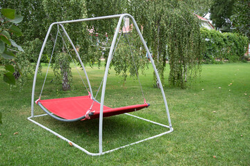 Red hammock swing in metal frame with nobody on green lawn in backyard. Rest relax relaxation alone on hammock swing in Summer garden.