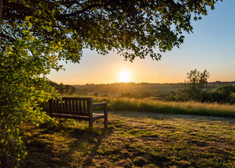 Lone bench seat overlooking countryside view with sunset orange sky nature reserve overhanging tree silhouette perfect summer sunset viewpoint peaceful fields forest woodland leaves sunlight no people - Powered by Adobe