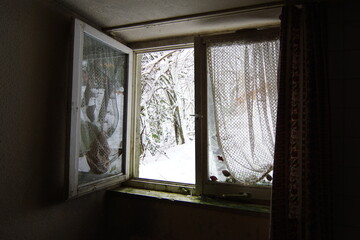 open window with old curtains in an abandoned house, outside there is snow	