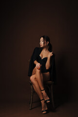 A young woman sits on a chair, in a jacket draped over her shoulders, against a solid dark brown...