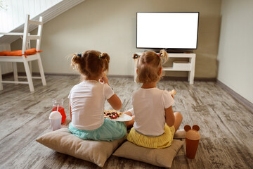 Two little girls are sitting on the floor on pillow, eating pizza and watching tv, playing games at home indoors. Two sisters spend time together, happy childhood.