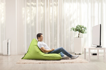Young man sitting on a green bean bag armchair and watching tv at home
