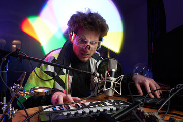 Angry caucasian composer looks at camera and plays music using midi keyboard in dark room with lights.