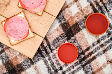 Sandwiches on the wooden stand with orange mugs from thermos on the plaid. Cozy picnic on fresh air.