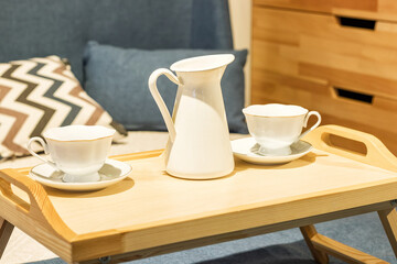 wooden tea table stands on the bed with cups. Bed with pillows and blue duvet and wooden chest of drawers