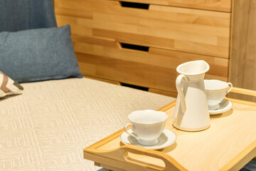 wooden tea table stands on the bed with cups. Bed with pillows and blue duvet and wooden chest of drawers